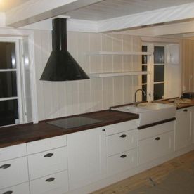 Et hvitt åpent kjøkken med et landlig preg på skap og skuffer som er hvitmalt, en hvit keramikk kjøkkenvask, moderne inventar, naturtre benkeplate og hvite trevegger