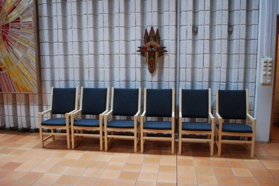 Stoler av lyst tre med rygg og sete av stoff plassert i en rad inne i en bygning med en skulptur som henger på veggen over de