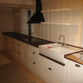 Et hvitt åpent kjøkken med et landlig preg på skap og skuffer som er hvitmalt, en hvit keramikk kjøkkenvask, moderne inventar, naturtre benkeplate og hvite trevegger