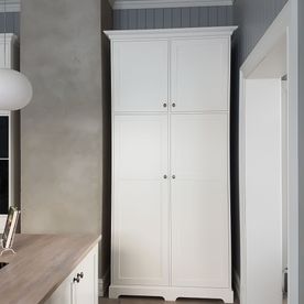 Et høyt hvitt kjøkkenskap i et mellomrom mellom en vegg og en skorsteinspipe som står i stil med resten av kjøkkenet